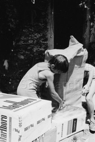 Bosco della Bella, Feriendorf für kinderreiche Familien - Zwei Kinder stülpen Jutesack über grosse Kisten mit "Marlboro" Aufdruck; um 1975