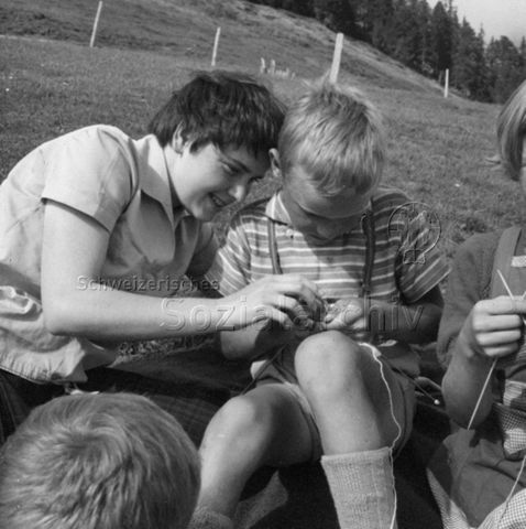 Praktikantinnenhilfe - "Die Buben wollten nicht zurückstehen. Mit Eifer lernen auch sie das 'Ine stäche - ume schloh'. Die Praktikantin übt sich im Anleiten von Kindern und die Mutter ist dankbar auch für diese Form von Hilfe"; um 1965