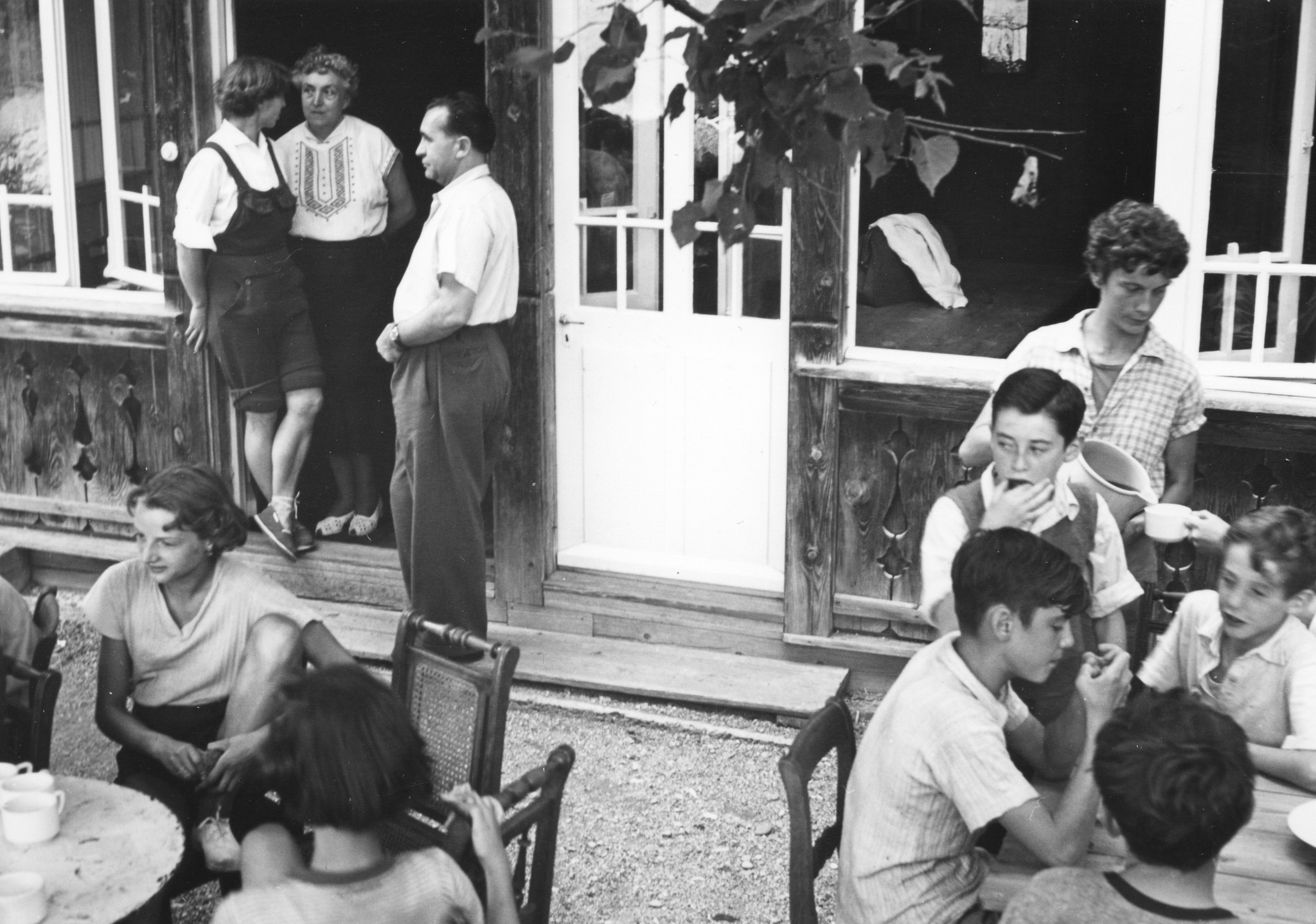 SAH Kinderlager im Sommer 1952 in Kienthal - Kinder sitzend draussen an Tischen und trinken Tee; drei Betreuer sprechen an der Tür miteinander