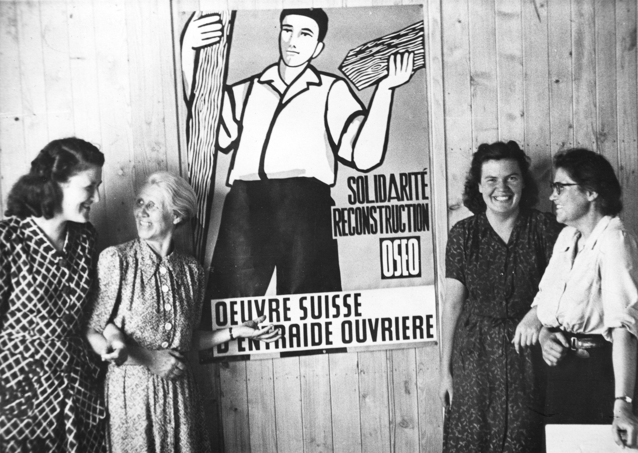 "Fives [Stadtviertel von Lille, Nord-Pas-de-Calais]", lachende Frauen neben einer SAH-Plakate mit Bauarbeiter: "Solidarité, Reconstruction, OSEO, Oeuvre suisse d'entraide ouvrière", um 1946