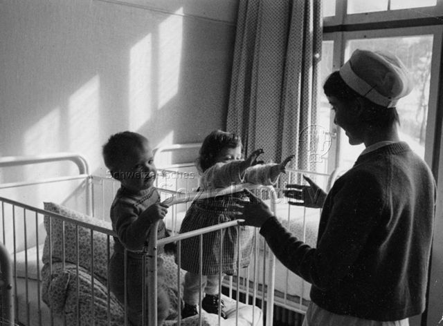 "Heimkinder" - Zwei Kleinkinder stehen im Gitterbett, die Betreuerin steht vor ihnen; um 1970