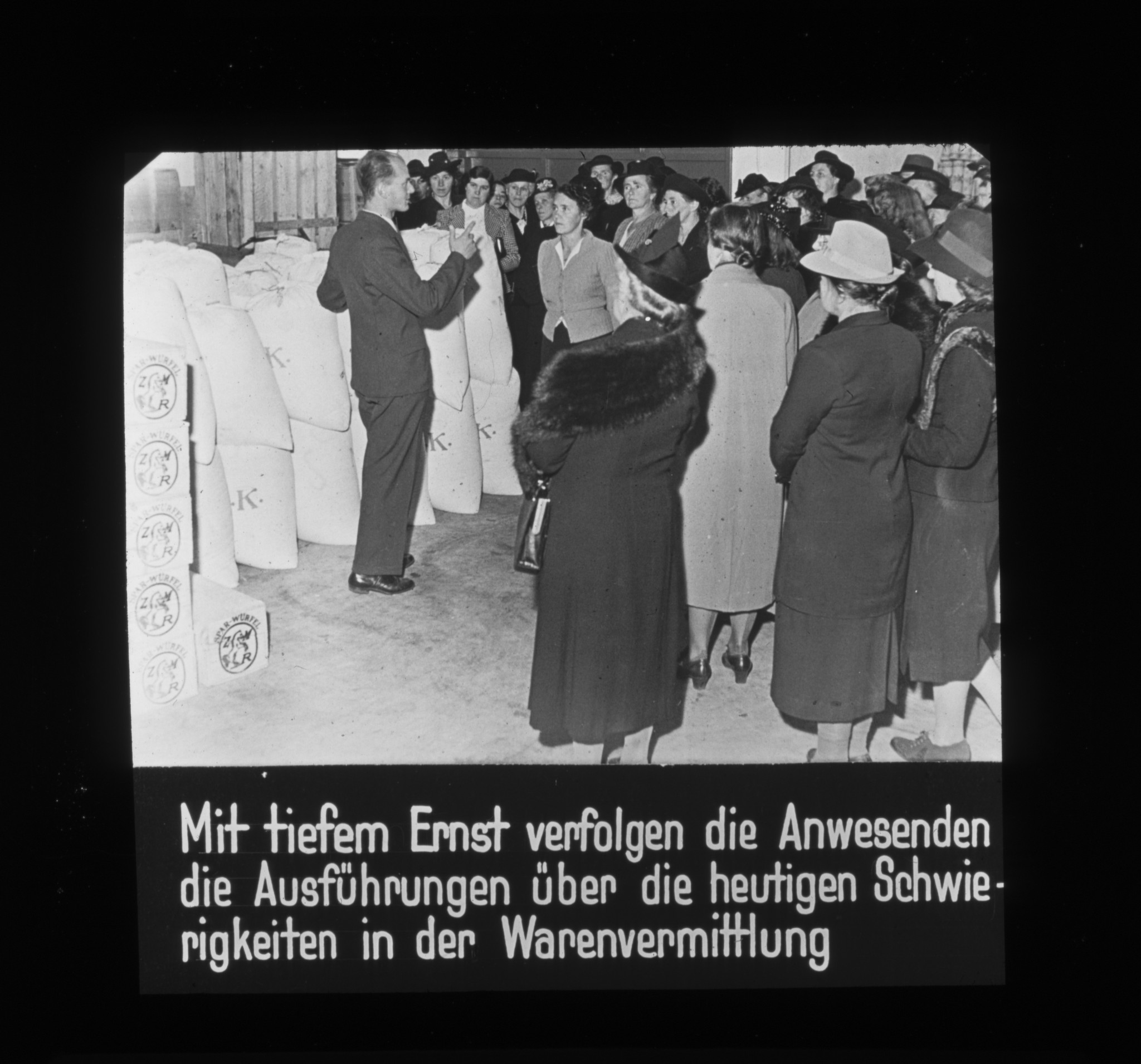 Dia: "Mit tiefem Ernst verfolgen die Anwesenden die Ausführungen über die heutigen Schwierigkeiten in der Warenvermittlung". Mann neben Säcken beim Sprechen vor einer Gruppe von Frauen