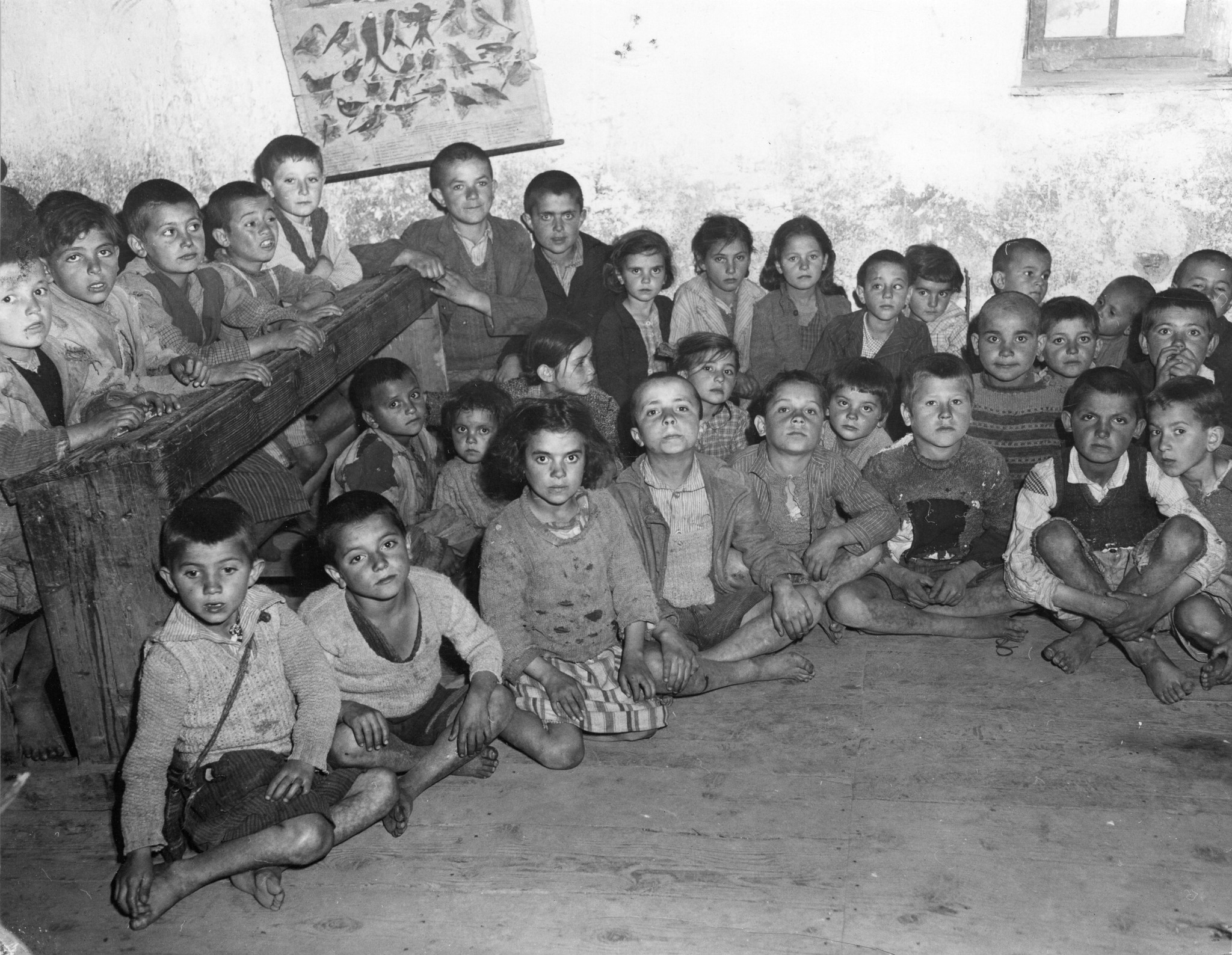 "Griechenland 1950. Ein Schulzimmer. Die Kinder sitzen auf einem Holzscheit oder am Boden, weil es nicht genügend Bänke hat."