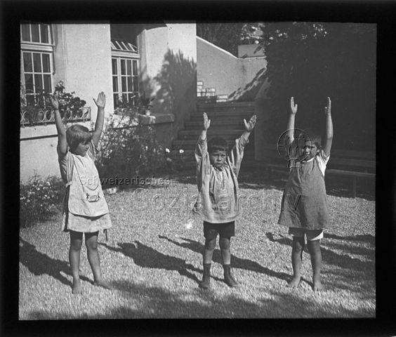 Diaserie zum Thema Kindergarten / Schulkind; drei Kinder mit erhobenen Händen auf einem Kiesplatz vor einem Haus stehend ; um 1930