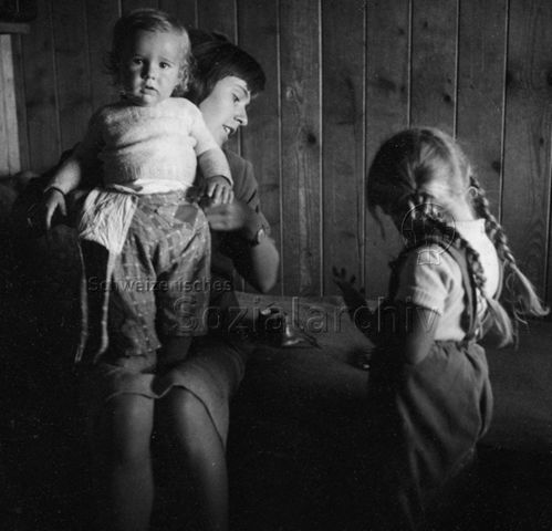 Praktikantinnenhilfe - Praktikantin mit Kleinkind auf dem Schoss, ein Mädchen steht daneben; um 1965