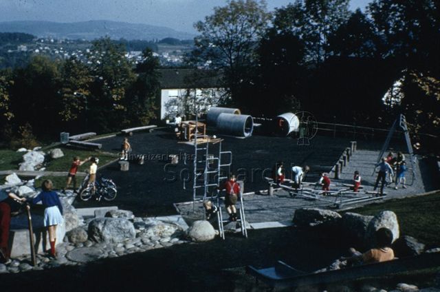 "Siedlungs- und Quartierspielplätze: Siedlungsspielplatz Adliswil ZH" - Blick auf den Spielplatz, Kinder spielenbei der Rutsche, auf den Schaukeln, auf dem Hartplatz; um 1970