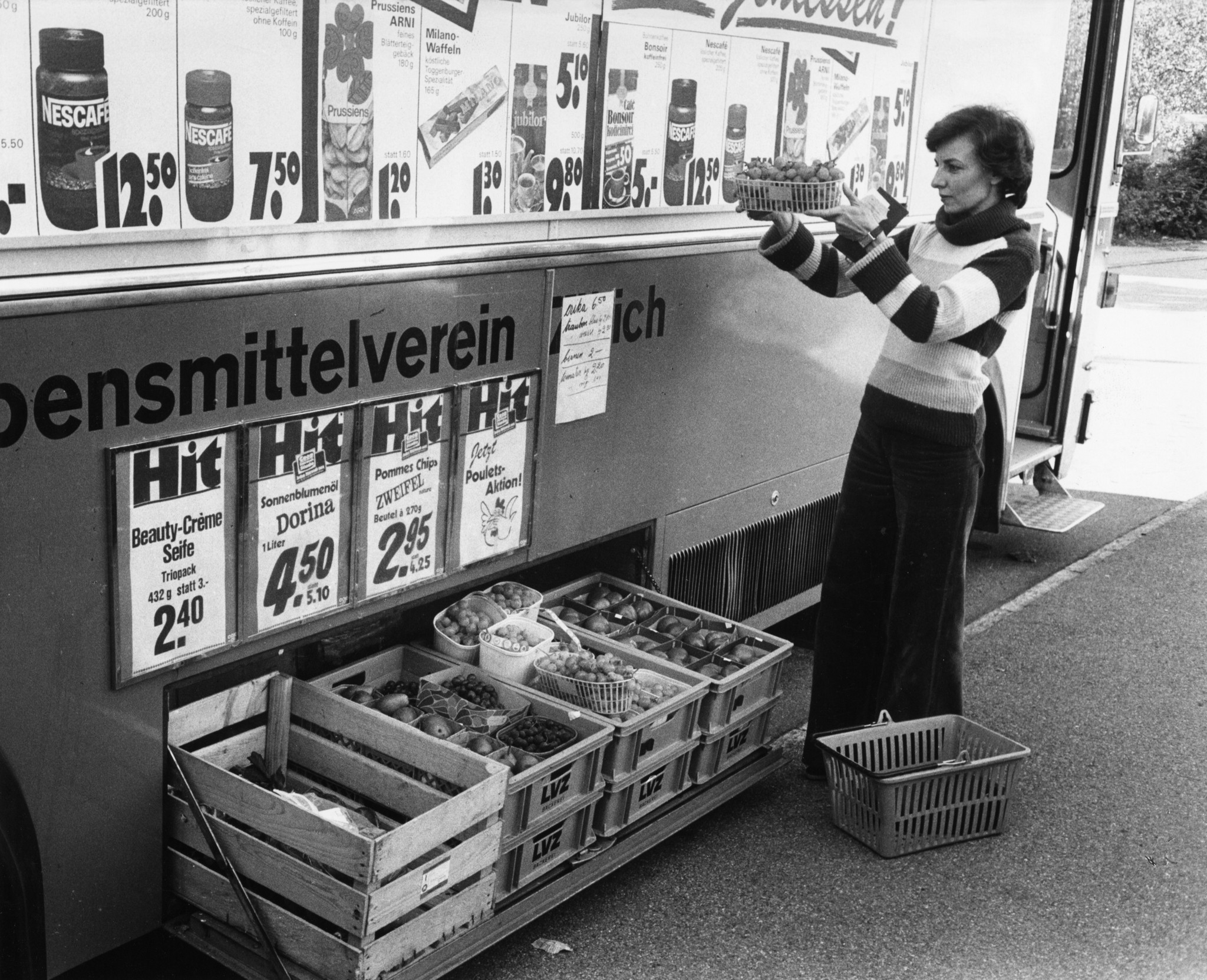 Kundin beim Begutachten eines Früchtekorbes vor einem Verkaufswagen des Lebensmittelvereins Zürich, weitere Kisten mit Ware und Werbetafeln