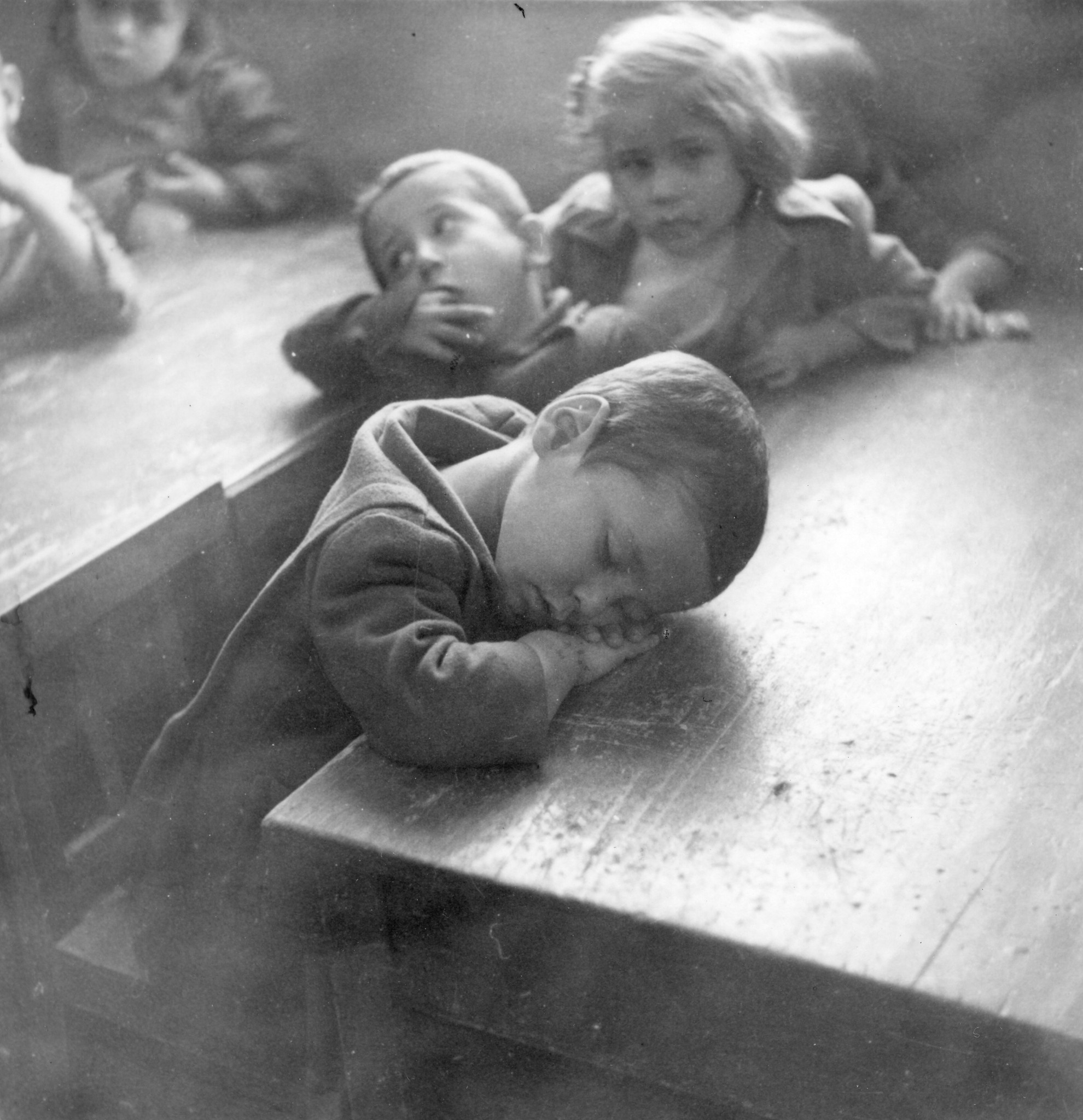 Kinder sitzen an einem Tisch; ein Knabe schläft mit dem Kopf auf dem Tisch (Schule?)