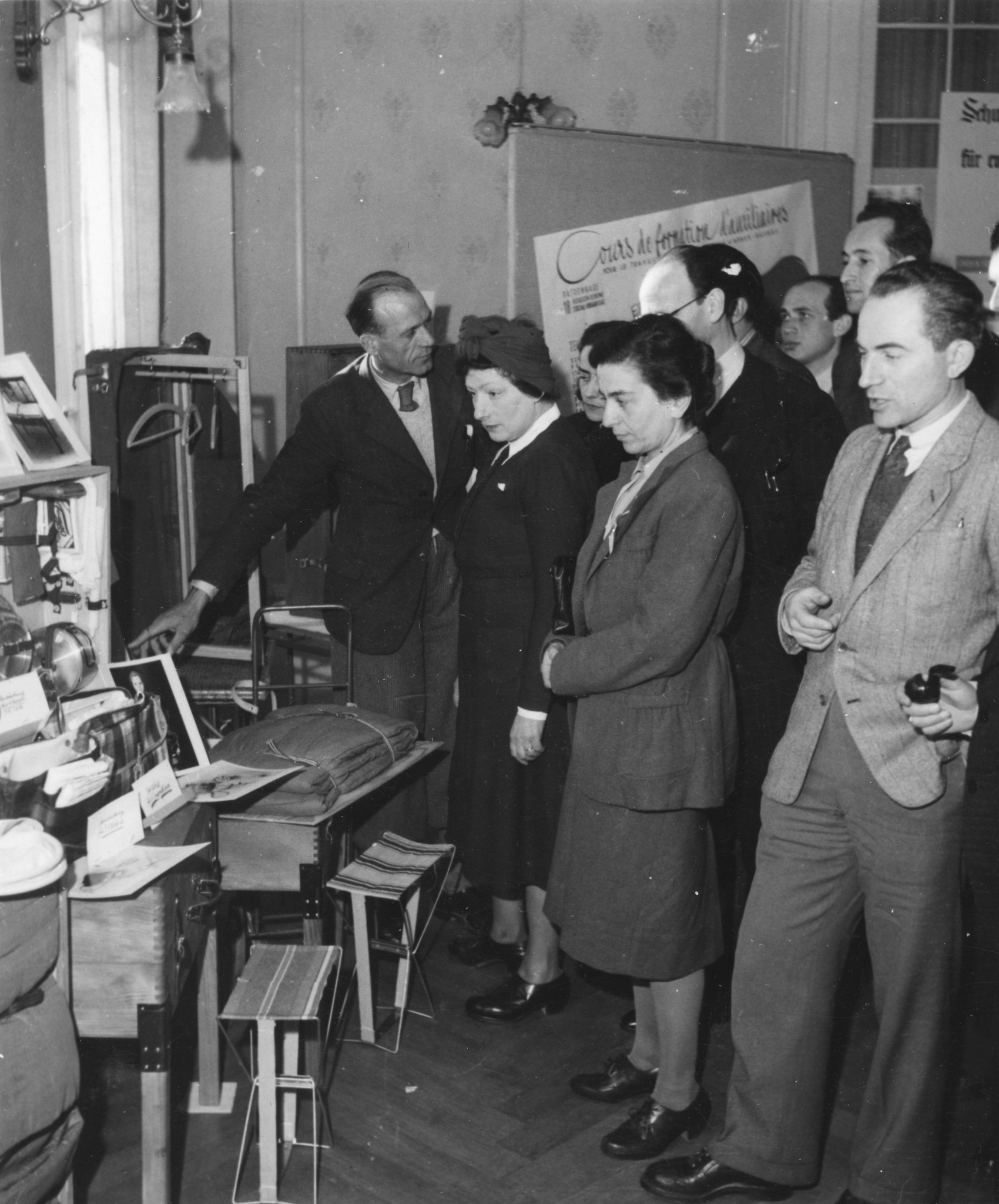 "Flüchtlingskongress, Montreux, Februar 1945. Ausstellung des SAH von Flüchtlings-Ausstattungsmaterial. Erfinder: Knellesen", Gäste besuchen eine Ausstellung mit Hilfsgütern (Küchenausstattung, Decken, Kleidungskoffer, Hocker...)