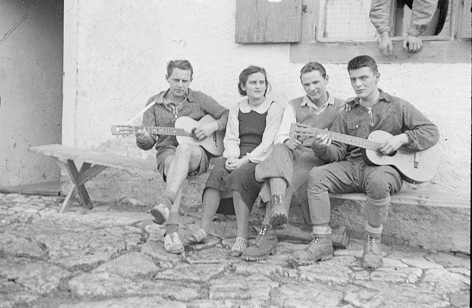 Vier Wandervögel sitzen auf einer Holzbank vor einem Haus; zwei spielen Gitarre