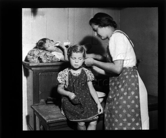 Diaserie "Reserve" und "alte Dias"; eine Frau am Flechten der Haare eines Mädchens; im Hintergrund ein Mädchen mit einer Trinkflasche im Mund auf einem Holzschrank liegend; um 1950