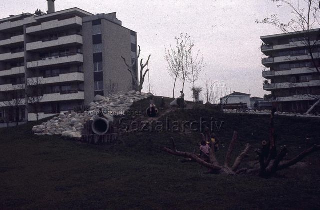 "Siedlungs- und Quartierspielplätze: Selbstbau Siedlungsspielplatz Moos, Oberhünenberg ZG, 1977" - Kinder auf Kletterbaum und Kriechrohren sitzend, zwischen Wohnblöcken