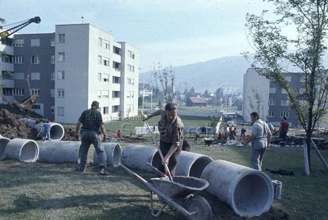 "Siedlungs- und Quartierspielplätze: Siedlungsspielplatz Stocken, Ebnat-Kappel SG" - Baustelle des Kinderspielplatzes auf einer grossen Wiese, Männer beim Auslegen von Rohren; um 1970