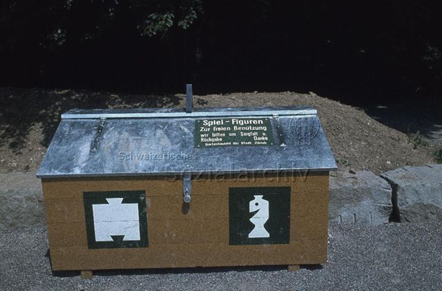 "Siedlungs- und Quartierspielplätze: Siedlungsspielplatz Buchholz/ Wittikon, Zürich" - Grosse Holztruhe im Freien mit Aufschrift "Spiel-Figuren zur freien Benützung" der Stadt Zürich; um 1970