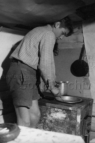 Junge beim Kochen mit einem Holzofen, wahrscheinlich in einer Spielplatz-Hütte; um 1965