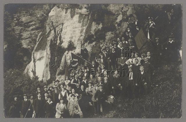 Gruppenporträt der Sozialistischen Jugendorganisation S.J.O. Freie Jugend mit Instrumenten und Fahnen, während einer Sonntagstour der Sektionen Luzern, Kriens, Emmenbrücke und Perlen-Root ins Melchtal, im Hintergrund Fels mit unleserlicher eingravierter Schrift, um 1919