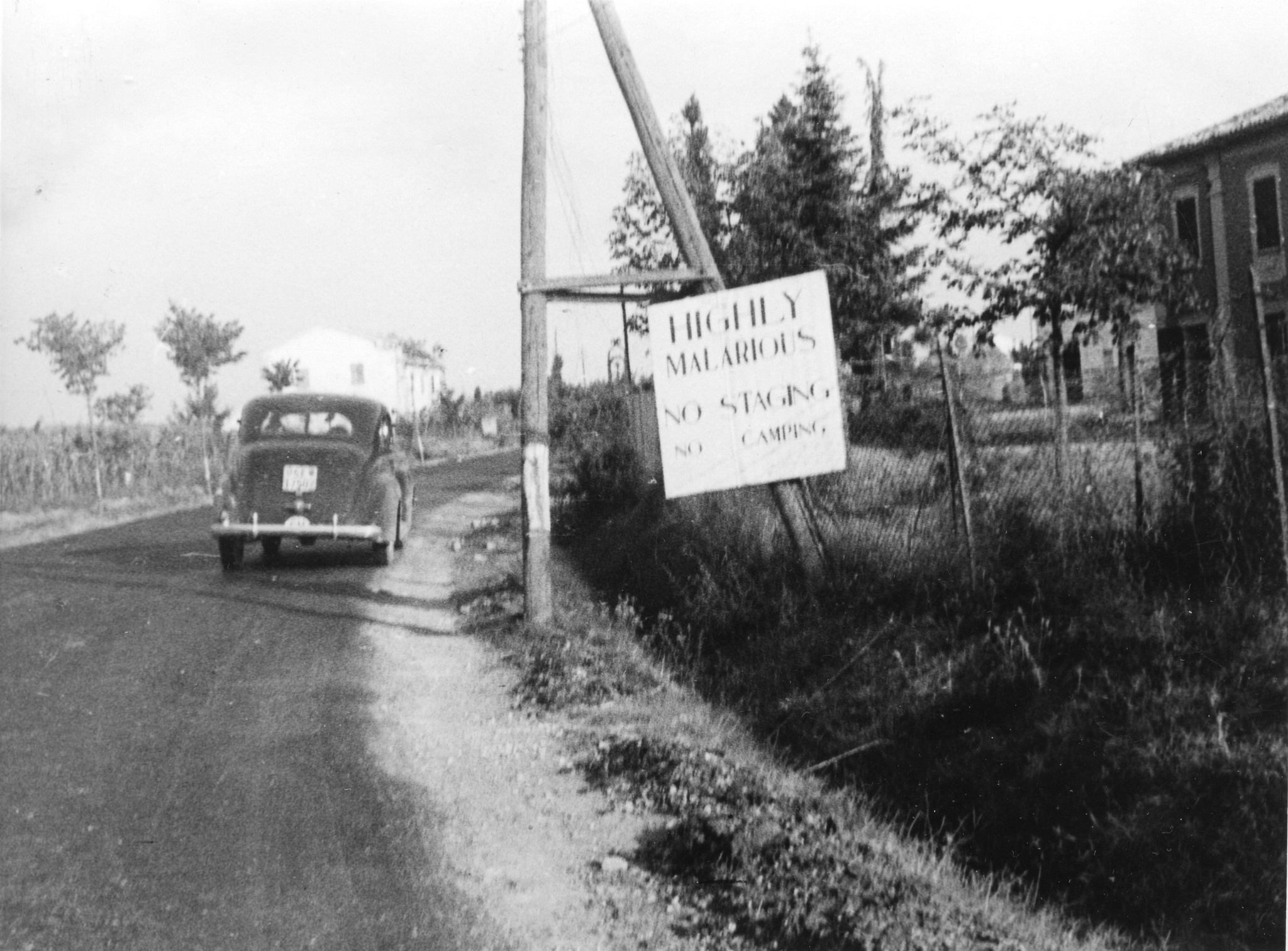 "In Emilien. Die Inschrift besagt auf Englisch: Achtung! Virulente Malaria. Nicht stehen bleiben, nicht campieren!"; Auto fährt auf einer Strasse, am Strassenrand steht ein Schild