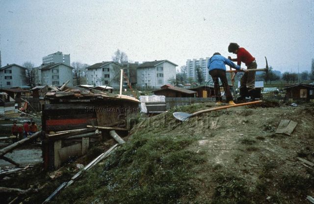 Robinsonspielplatz "Zug" - Kinder graben ein Loch, dahinter selbstgebaute Holzhütten; um 1975