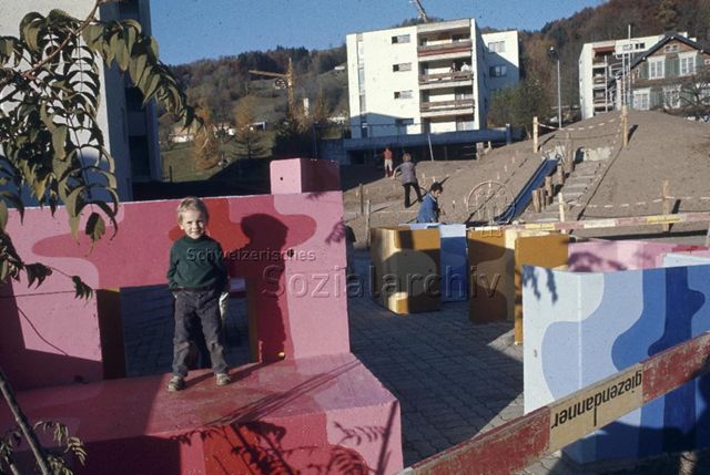 "Siedlungs- und Quartierspielplätze: Siedlungsspielplatz Stocken, Ebnat-Kappel SG" - Ein Kind steht auf bunt bemalten Bauteilen auf Hartplatz, im Hintergrund sieht man Erwachsene beim Gärtnern; um 1970