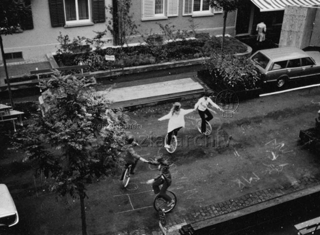 "Wohnen beginnt auf der Quartierstrasse" - Vier Kinder beim Einradfahren auf der Strasse; um 1975