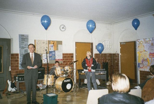 "Vernissage Plakate Aktionstag 1996" - Mann spricht zu Anwesenden, im Hintergrund wartet die Schülerband