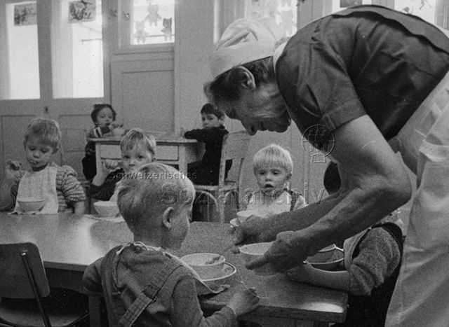 "Kinderheilstätte [Davos], Essen" - Kinder am Tisch, Krankenschwester beim Schöpfen von Essen; 1971