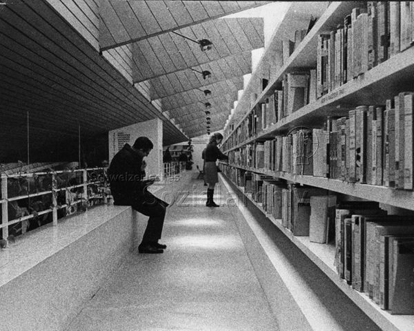 "Bibliothek Heuried" - Mann beim Lesen, Frau stöbert im Bücherregal; um 1970