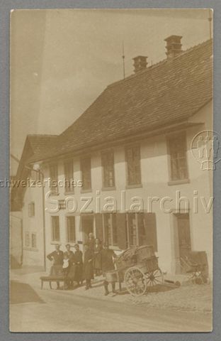 Uniformierter Postbote mit Paketanhänger, Familie und Kinderwagen vor Gebäude der "Post & Telegraph Rorbas-Freienstein