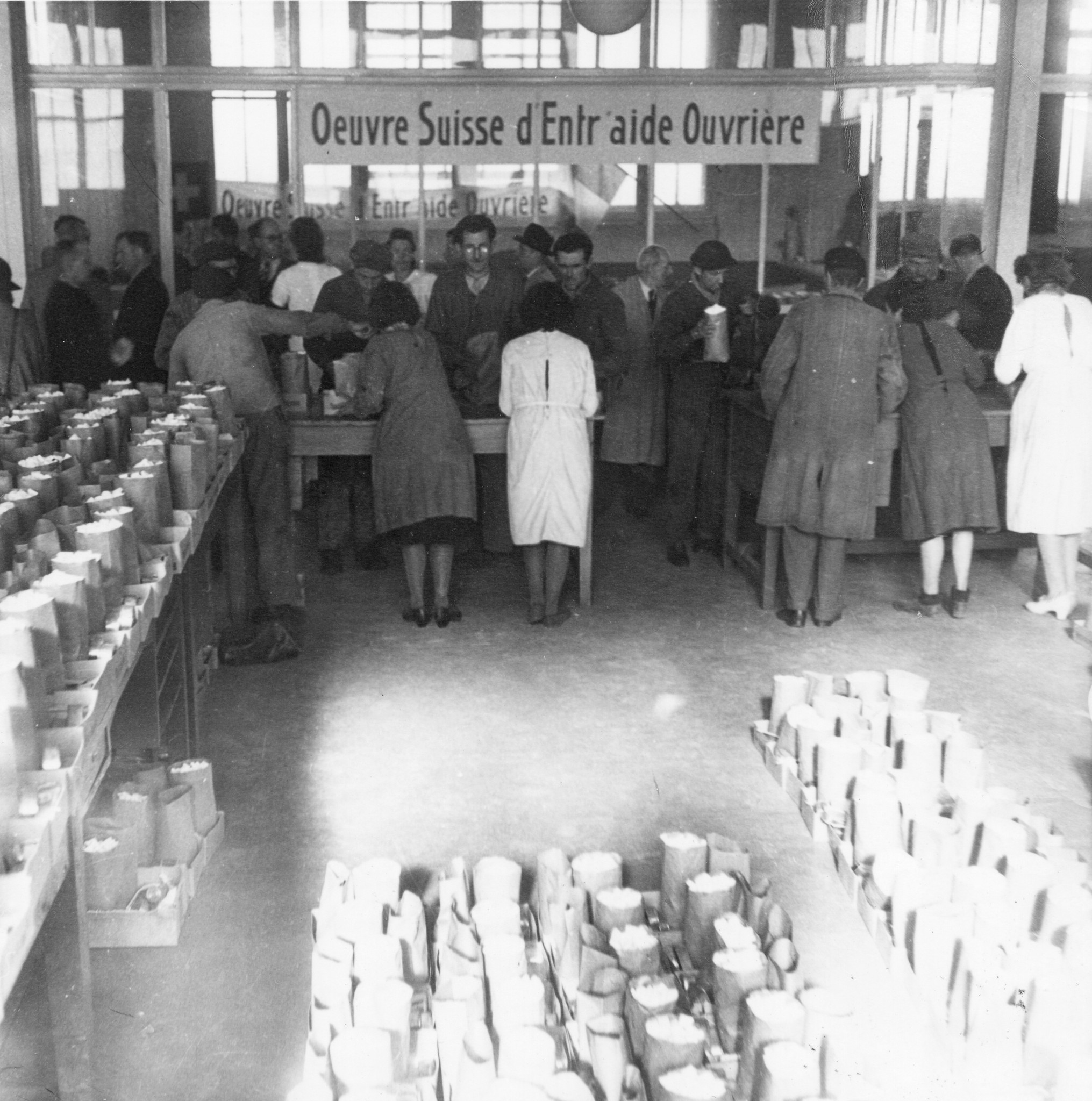 "Lyon, Oktober 1944. Die Verteilung beginnt Dienstag, den 3. Oktober 1944."  "Colis suisse', Jeder geschädigte Arbeiter erhält 2 kg. Zucker, 1 kg. Linsen, 1/2 kg. Teigwaren, 1 Büchse Tomatenpüree, 2 Büchsen Sardinen." Transparent: "OSEO"