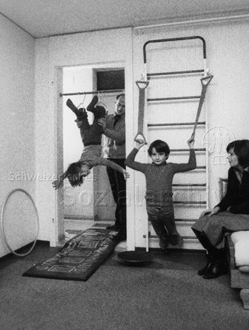 Familie beim Turnen - Das Mädchen hängt kopfüber im Türrahmen, der Vater hält sie fest, der Junge macht Übungen mit Gummibändern und die Mutter sitzt daneben; um 1970