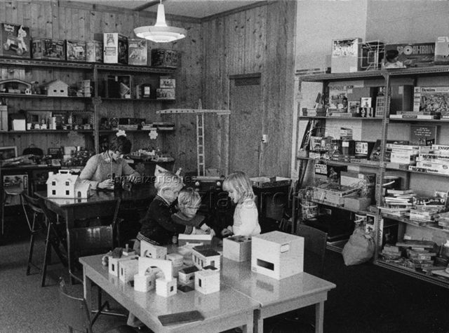 "Ludothek Nyon" - Drei Kinder beim Spielen, hinten sitzt ein Mann mit einem Spielzeug am Tisch; um 1970