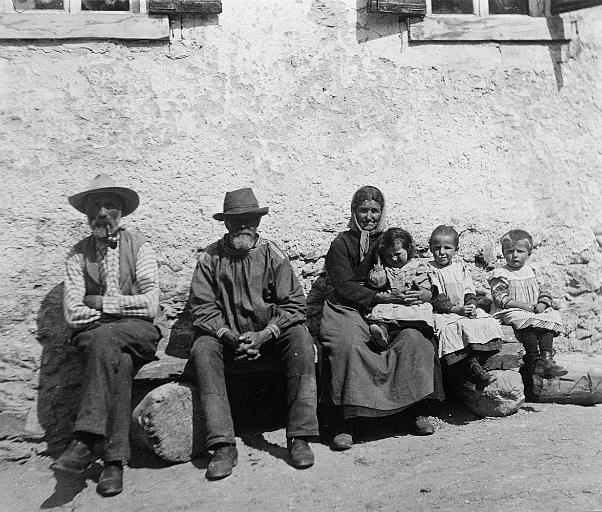 Von einer Bündnerfahrt - Drei Erwachsene und drei Kinder sitzen auf einer Holzbank vor einem Haus (1913)