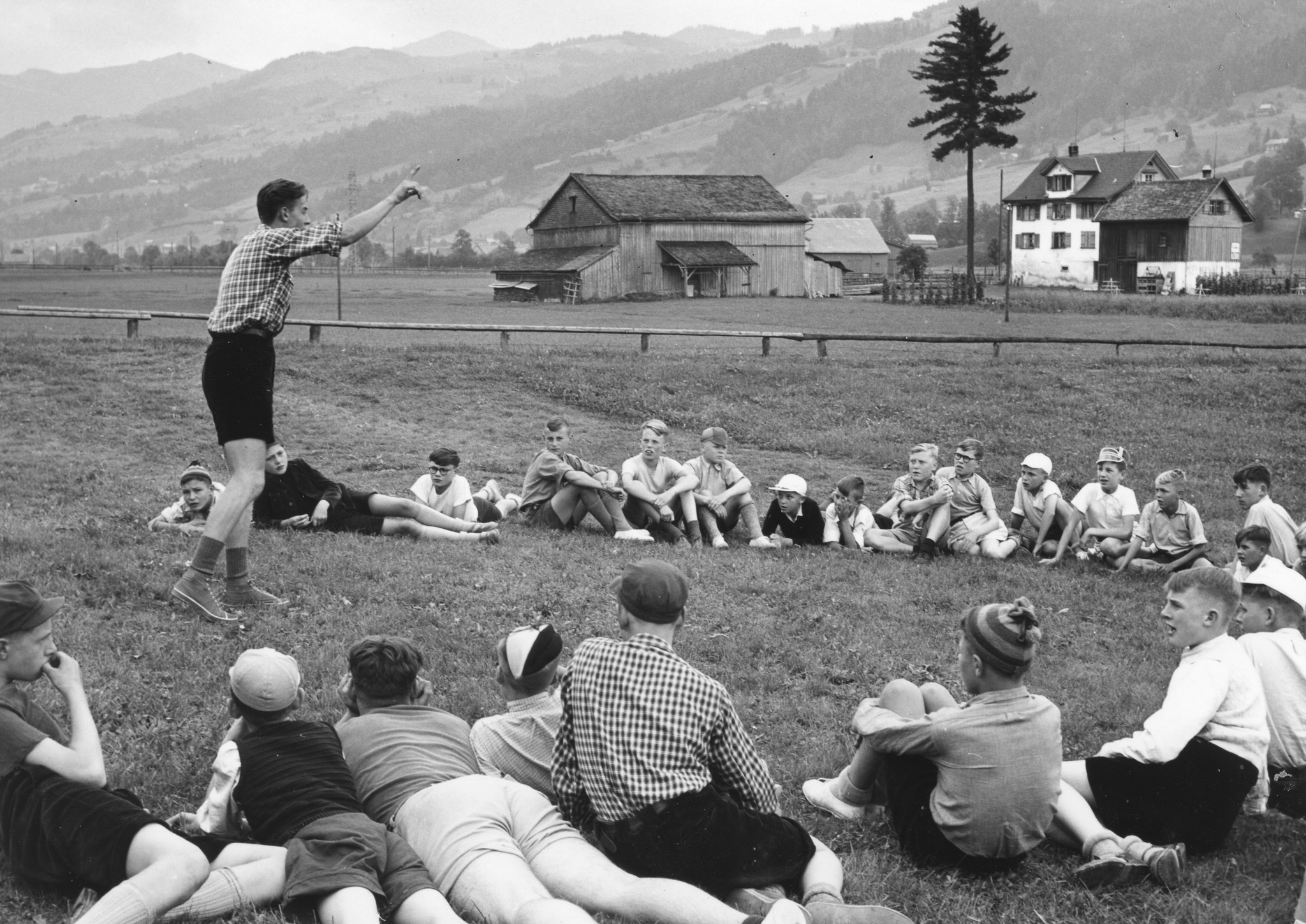 "Drei Eidgenossen", Wattwil. Belgierkolonie, Sommer 1950"; Kinder sitzen im Kreis auf einer Wiese und hören einem Betreuer zu