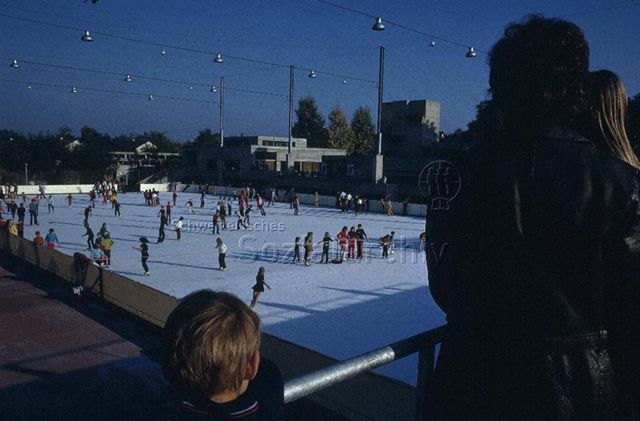 "Freizeitanlagen, Gemeinschaftszentren: Eisfeld FZA Heuried, Zürich" - Kinder am Eislaufen, Eltern und ein Kind schauen zu; um 1970