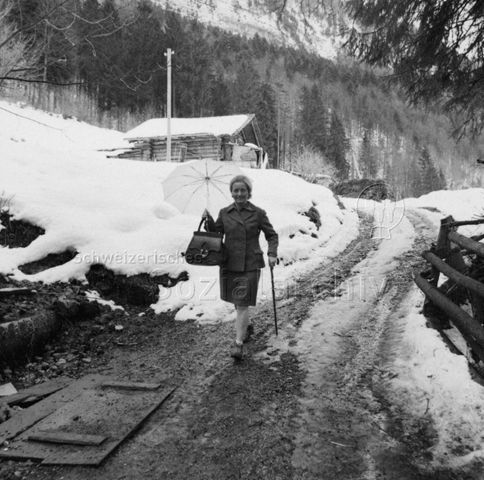 Frau geht mit Gehstock und Schirm über einen Feldweg, Winter; um 1965