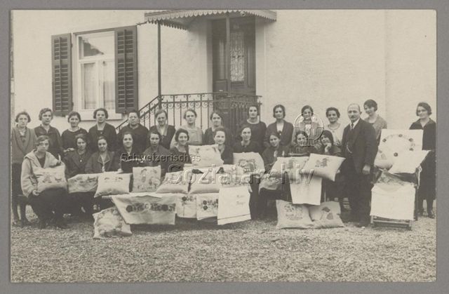 Gruppenporträt junger Frauen und einem Mann mit ihren Stickereiwaren (?) mit diversen Motiven, wahrscheinlich im Rahmen von Haushaltungsschule, um 1940