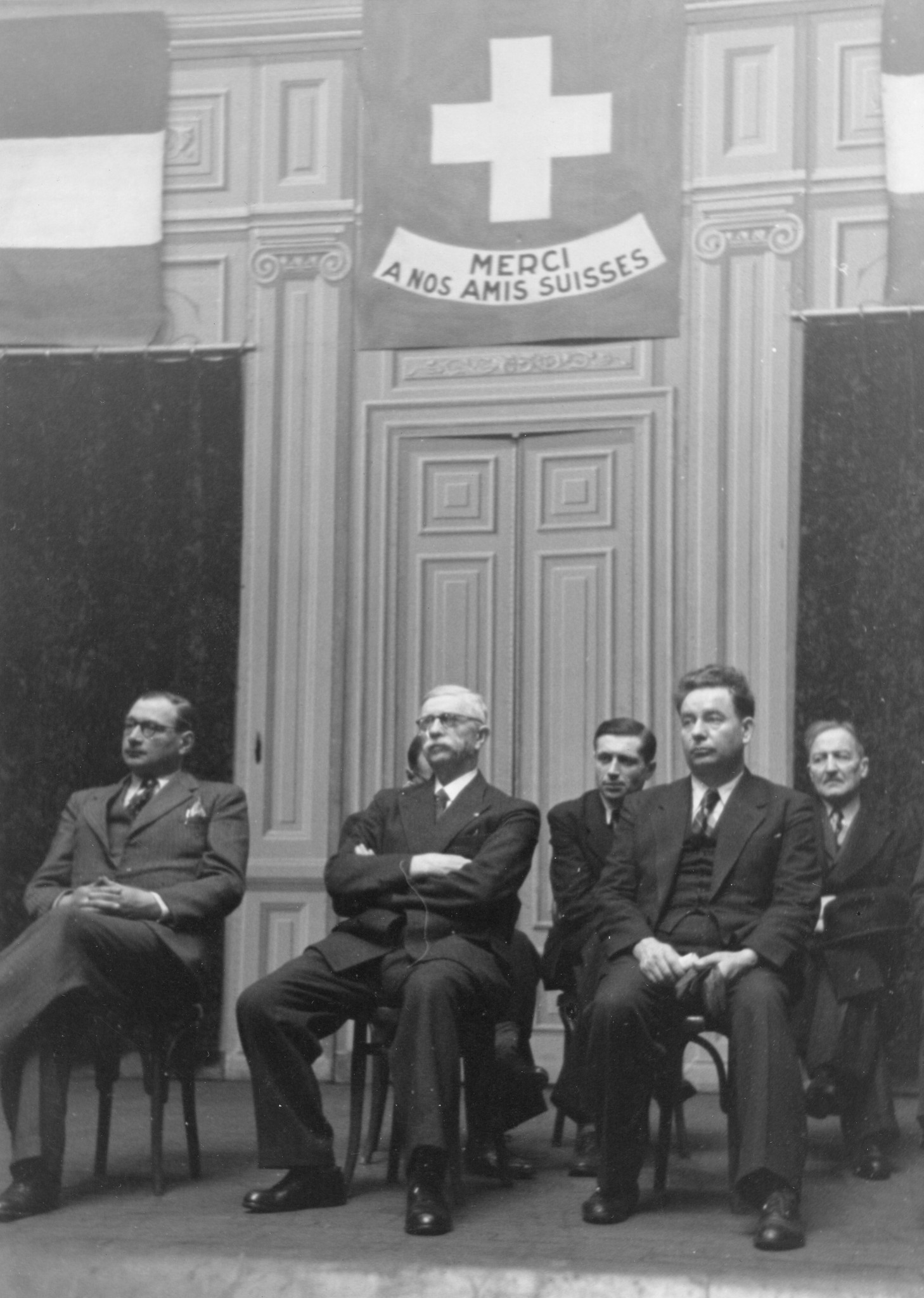 "Saint-Etienne, 26. Mai 1945. Jahrestagfeier der Bombardierung in der Bourse du Travail." Schweizer Fahne mit der Beschriftung: "Merci à nos amis suisses", Amtsinhaber und Amtsinhaberin auf einer Bühne