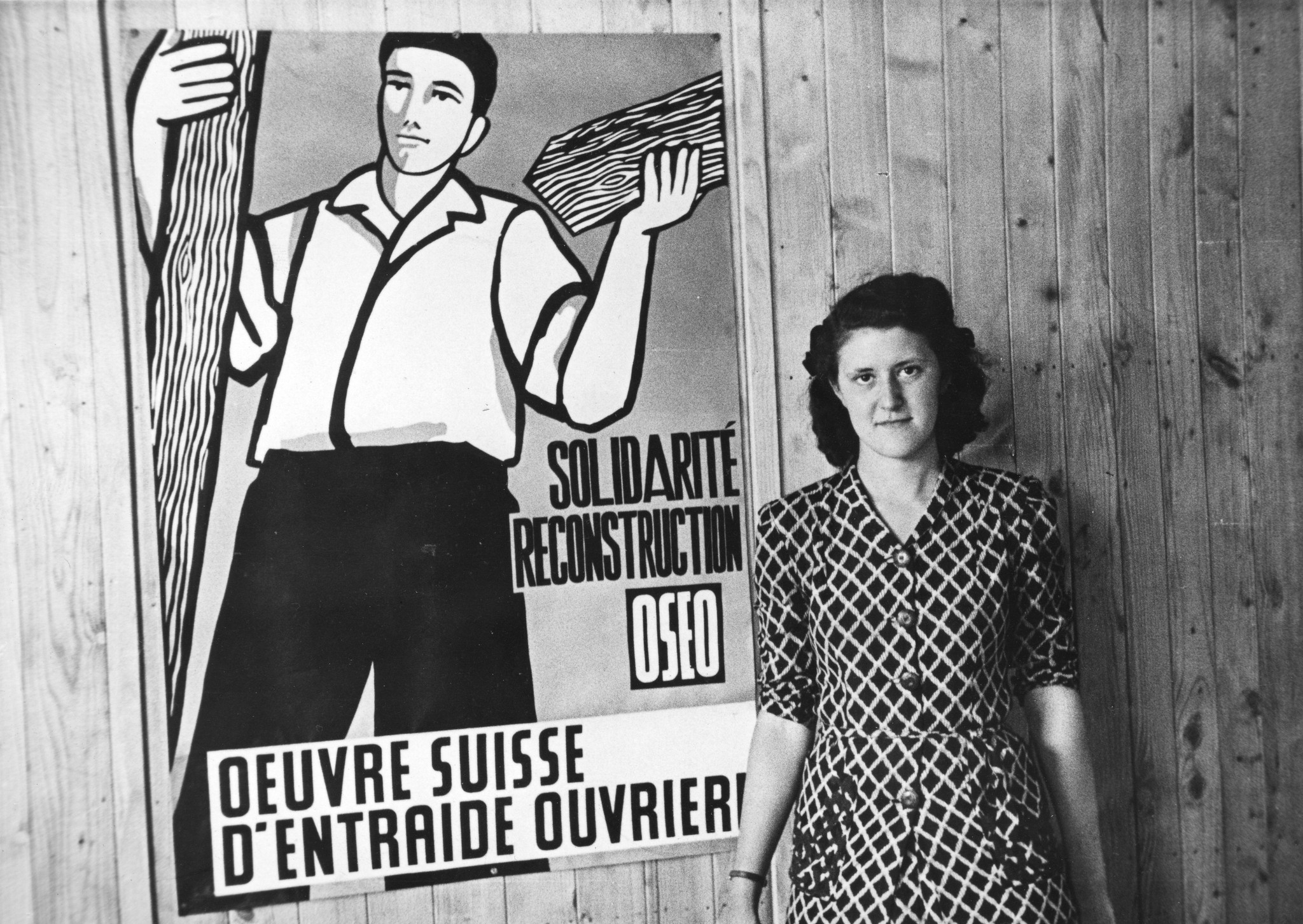 "Fives [Stadtviertel von Lille, Nord-Pas-de-Calais]", eine Frau neben einer SAH-Plakate mit Bauarbeiter: "Solidarité, Reconstruction, OSEO, Oeuvre suisse d'entraide ouvrière", um 1946