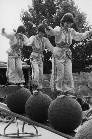 Drei Jugendliche bei einer Aufführung balancieren auf Bällen stehend über eine Wippe; um 1970