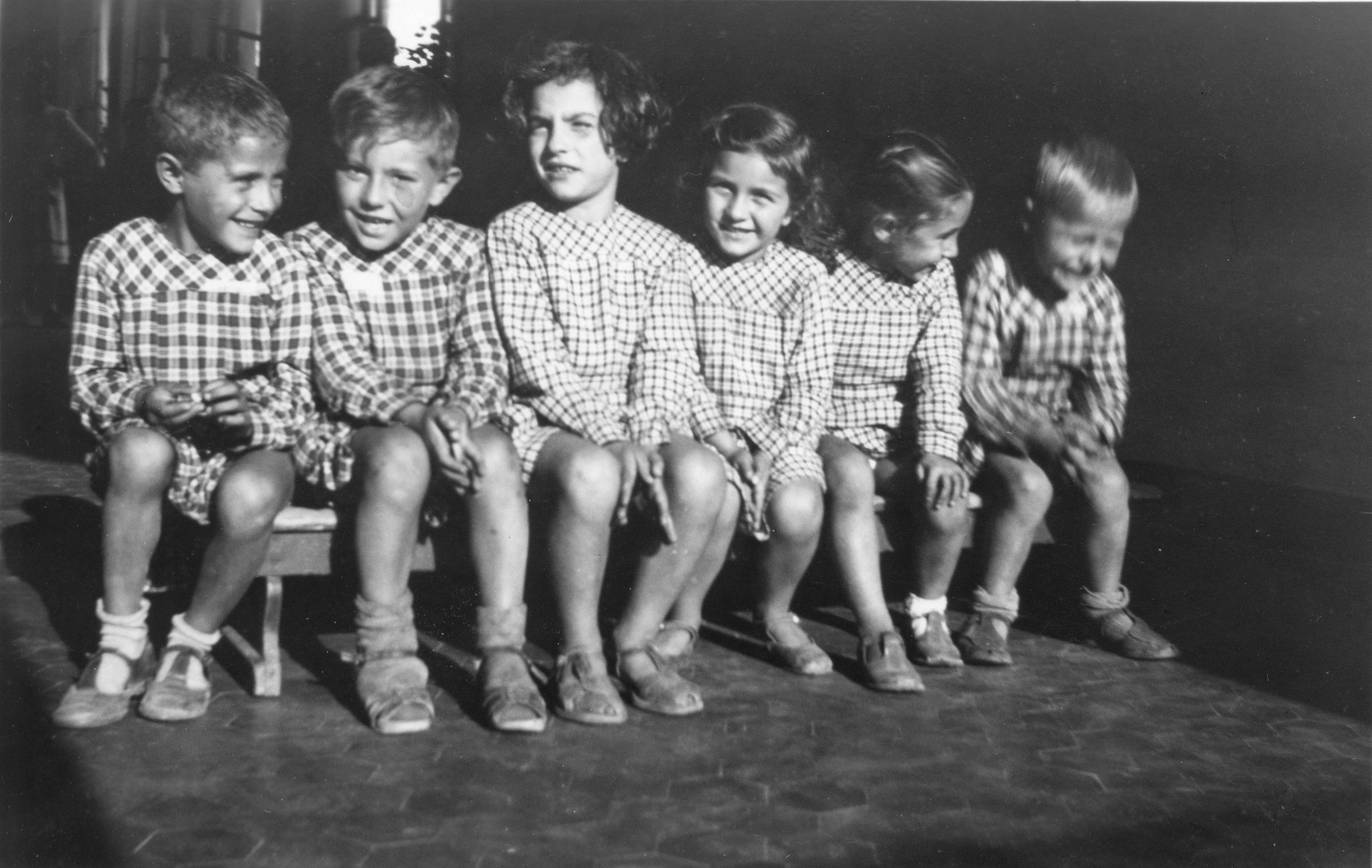 "Jetzt geht's nach Hause. - Die Mütter warten im Hintergrund. Wir benützen ein sonnenbeschienenes Plätzchen, um schnell ein Bild in unserer Halle zu machen."; Kinder sitzen auf einer Bank
