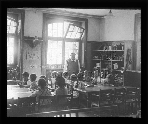Diaserie zum Thema Kindergarten / Schulkind; Kinder sitzen in einem Schulzimmer an zwei Tischen; Kinder am ersten Tisch weben mit Papier, Kinder am zweiten Tisch arbeiten mit Holzklötzen; Betreuerin am Kopfende des zweiten Tisches stehend; um 1930