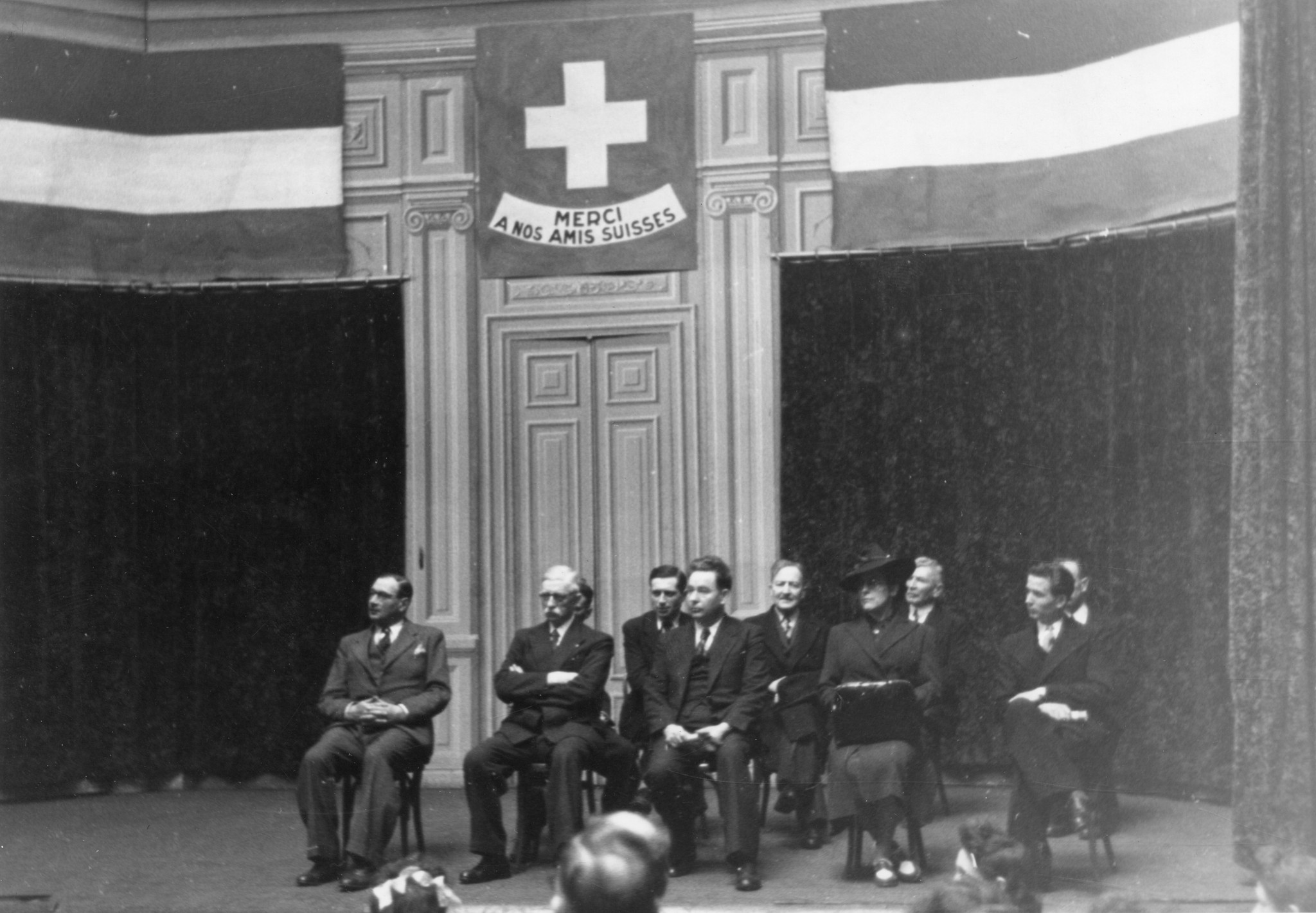 "Saint-Etienne, 26. Mai 1945. Jahrestagfeier der Bombardierung in der Bourse du Travail." Schweizer Fahne mit der Beschriftung: "Merci à nos amis suisses", Amtsinhaber und Amtsinhaberin auf einer Bühne