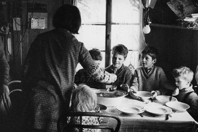 "Praktikantenhilfe" - Kinder sitzen am Tisch, Praktikantin schöpft Suppe; um 1965