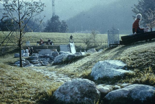 "Siedlungs- und Quartierspielplätze: Siedlungsspielplatz Adliswil ZH" - Blick in die grüne Umgebung, Kinder spielen auf Rutschbahn und auf den Steinen; um 1970