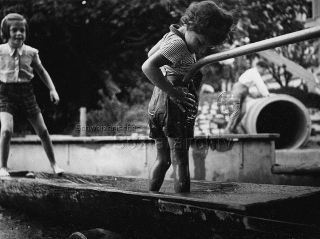 "Zug" - Kind steht im Brunnen und wäscht seine Hose; um 1960