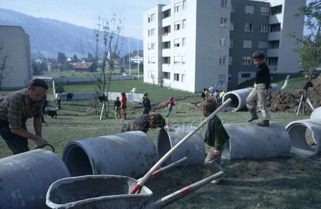 "Siedlungs- und Quartierspielplätze: Siedlungsspielplatz Stocken, Ebnat-Kappel SG" - Baustelle des Kinderspielplatzes auf einer grossen Wiese, Männer beim Auslegen von Rohren, Kinder spielen um sie herum; um 1970