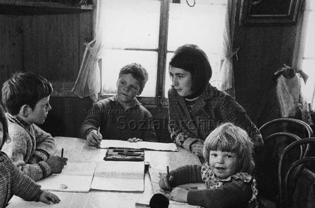 Praktikantenhilfe - Praktikantin hilft den Kindern bei den Schulaufgaben, ein Mädchen sitzt am Tisch und malt; um 1970
