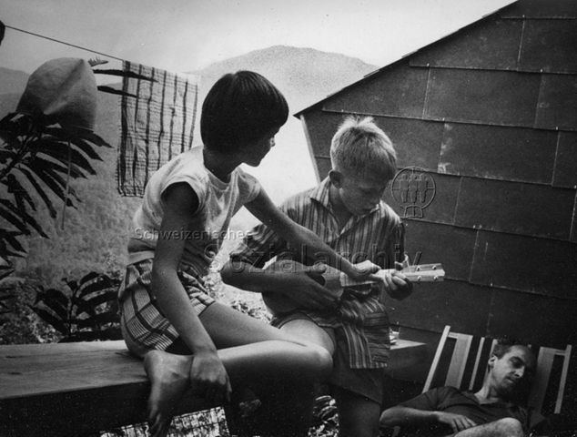 Bosco della Bella, Feriendorf für kinderreiche Familien - Auf dem Balkon, zwei Jungen spielen Ukulele, der Vater erholt sich im Liegestuhl; um 1970