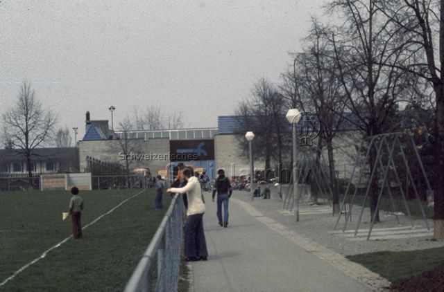 "Freizeitanlagen, Gemeinschaftszentren: FZA Heuried, Zürich, 1977" - Erwachsene und Kinder am Rande des Sportfeldes vor dem Betongebäude; um 1970
