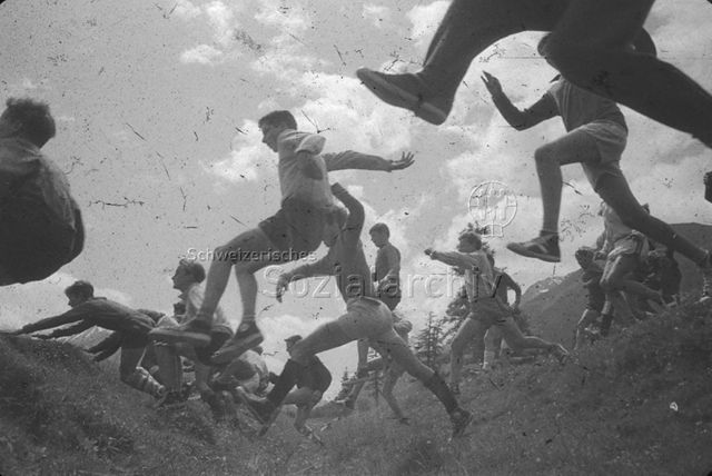 "Volkssport; Veranstaltungen: Bubenwettkampf" - Junge Männer in sportlicher Kleidung rennend durch eine Wiese, einige springen über eine kleine Senke; um 1960